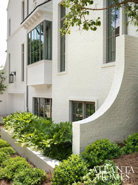White brick house with bay window, dark framed windows, and lush landscaping