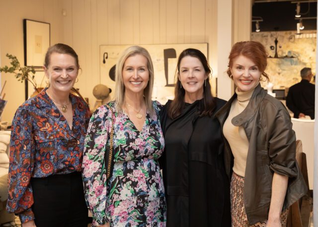 Four women smiling, two in floral dresses, one in a patterned blouse, one in a black dress and