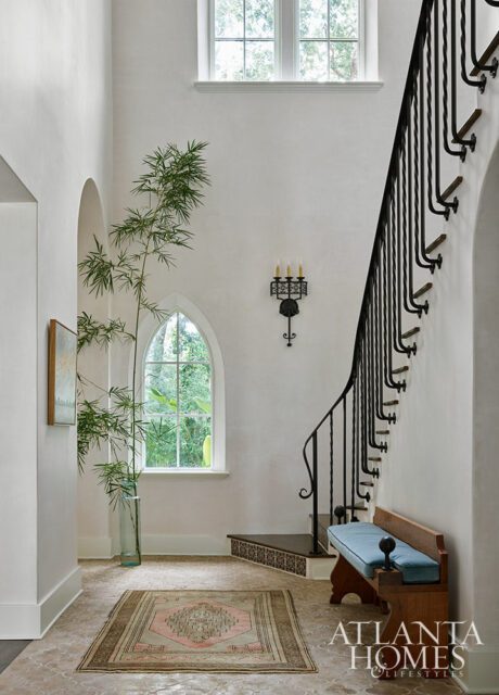 Hallway with arched window, bamboo plant, ornate staircase, and bench.
