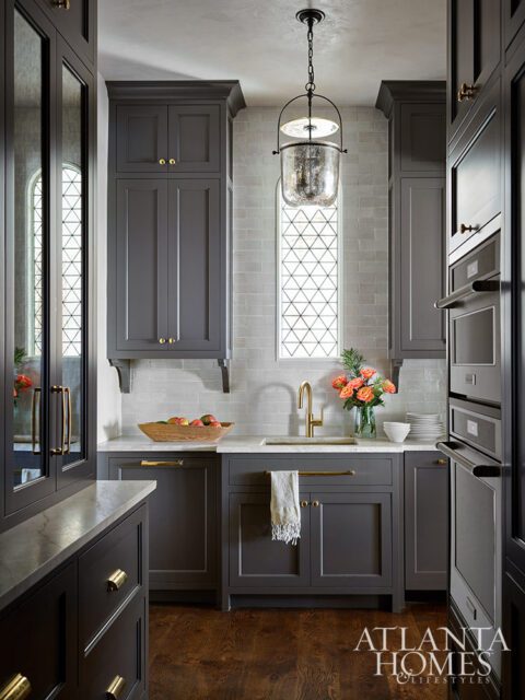 Gray kitchen cabinets with brass hardware, white subway tile backsplash, and a decorative window.