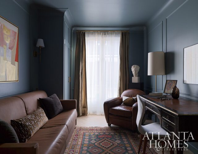 Dark blue room with brown leather sofa, armchair, and desk. Oriental rug on floor.