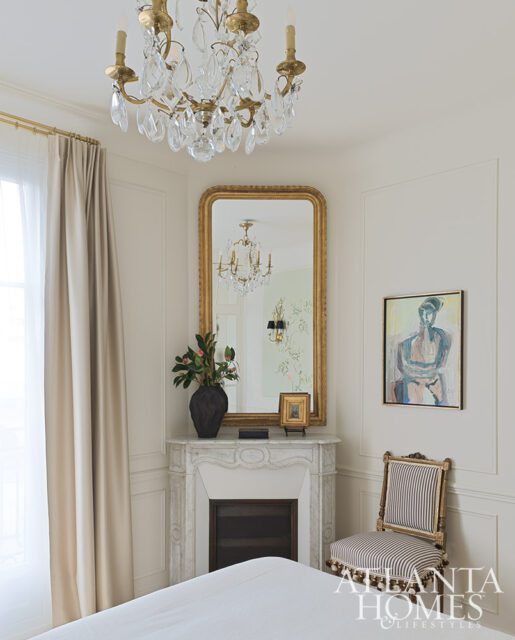 Bedroom with marble fireplace, gold mirror, crystal chandelier, and striped chair.