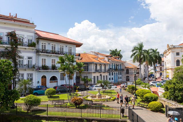 Light blue buildings with red tile roofs, balconies, and palm trees line a street with a fountain