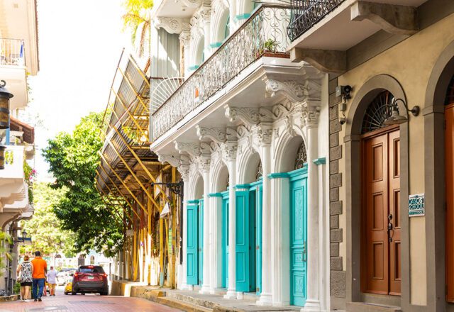 White building with ornate columns and turquoise doors, next to a tan building with brown doors.