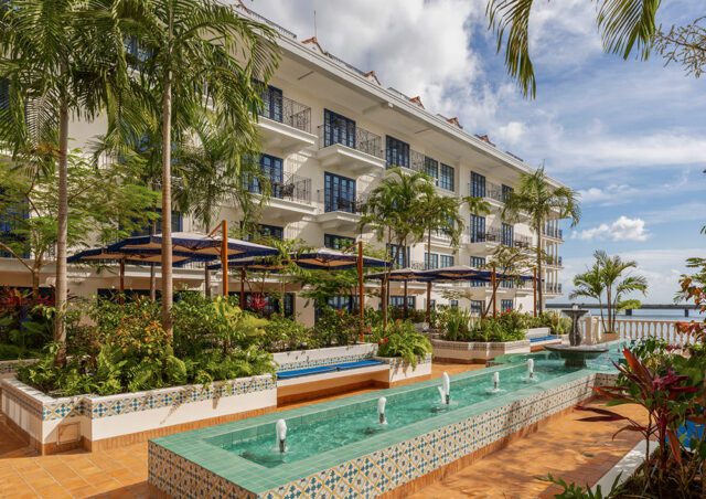 White hotel with balconies, palm trees, and a long water fountain feature.