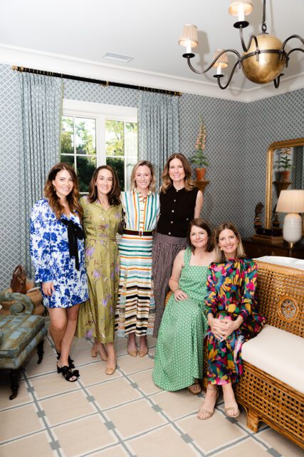 Six women posing in a room with blue patterned wallpaper, patterned curtains, and a rattan sofa.