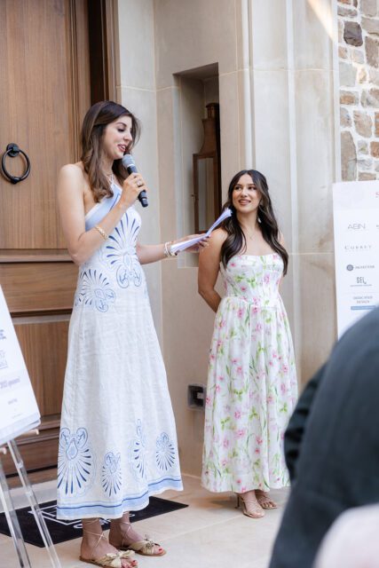 Woman in white dress with blue pattern speaks into microphone, woman in floral dress stands beside