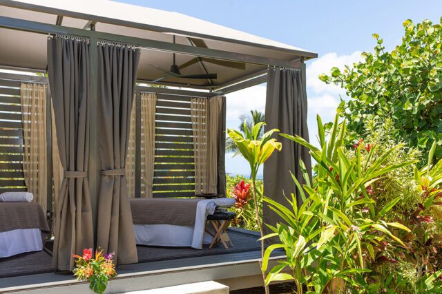 Outdoor spa cabana with massage tables, gray curtains, and lush tropical foliage.
