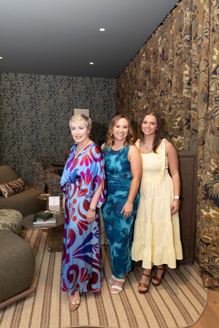 Three women posing in a room with patterned wallpaper and floral curtains.
