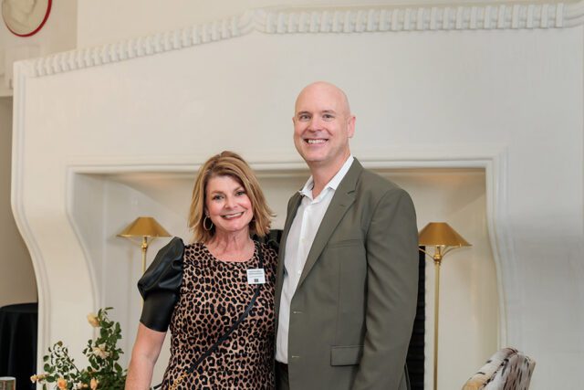 Woman in leopard print dress and man in olive suit smiling in front of fireplace
