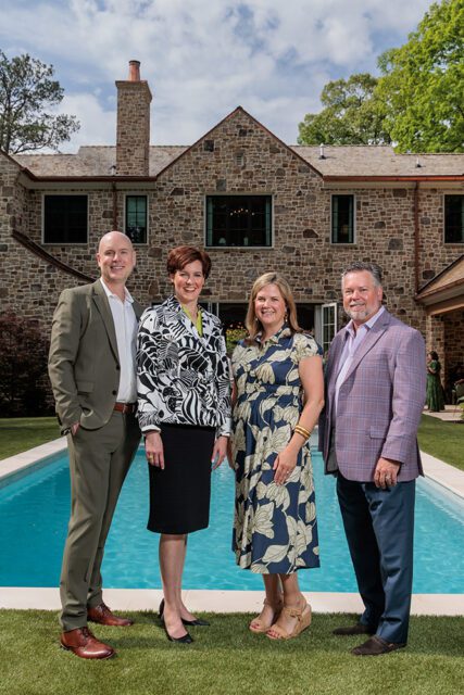 Four people standing in front of a stone house with a swimming pool