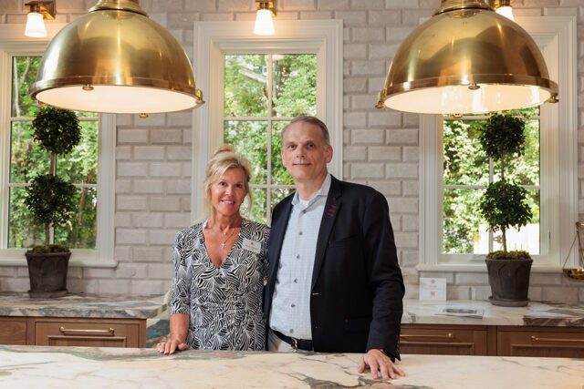 Woman in patterned dress and man in suit stand in kitchen with brass pendant lights and subway tile