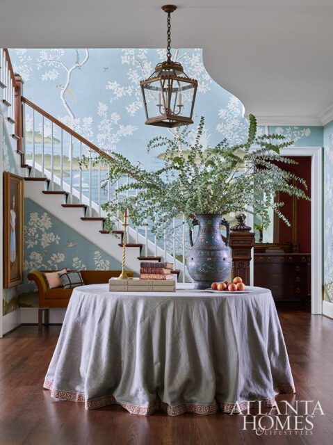 Foyer with blue chinoiserie wallpaper, staircase, round table with vase and books.