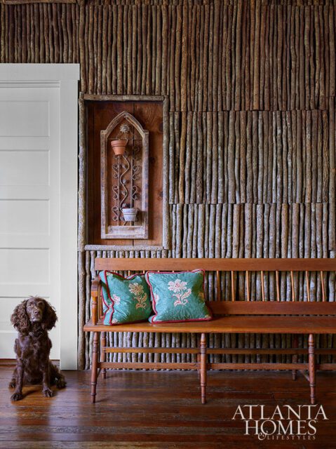 Wooden bench with floral pillows against a wall of vertical sticks. Dog sits nearby.