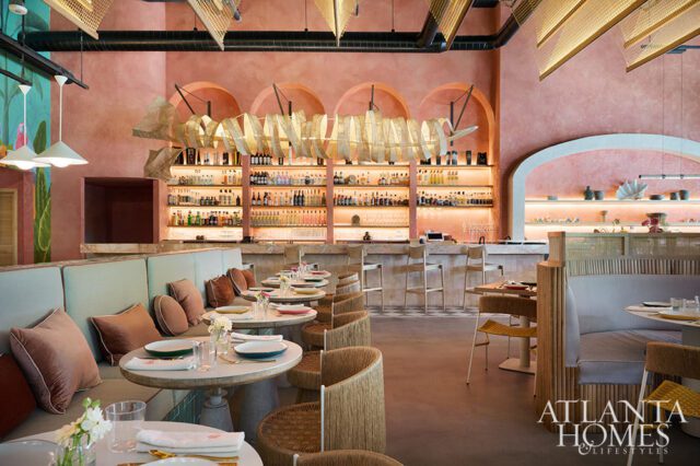 Restaurant interior with pink walls, light blue banquettes, rattan chairs, and a bar with shelves