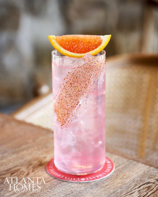 Pink cocktail with orange slice garnish and spiced rim, on coaster