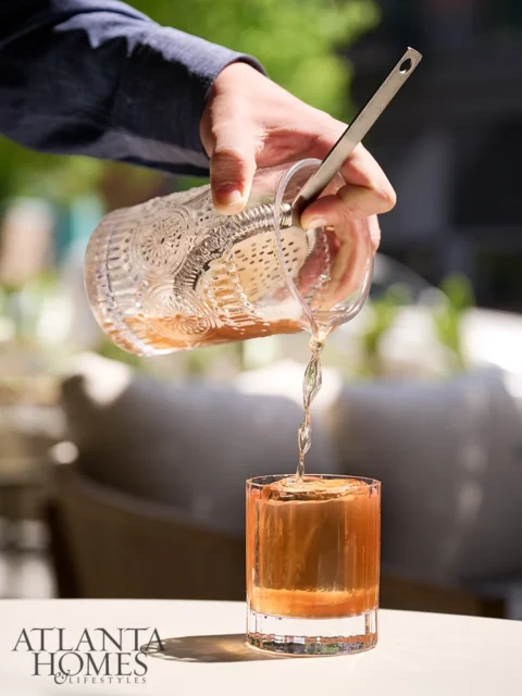 Pouring amber liquid from ornate glass pitcher into glass with ice