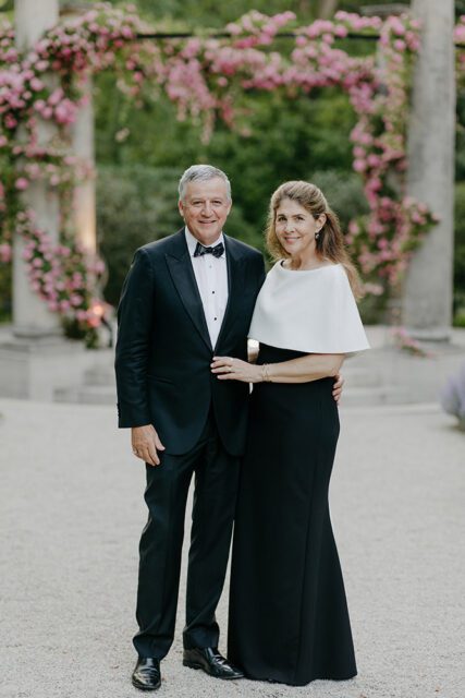 Man in tuxedo and woman in black dress with white cape stand outdoors