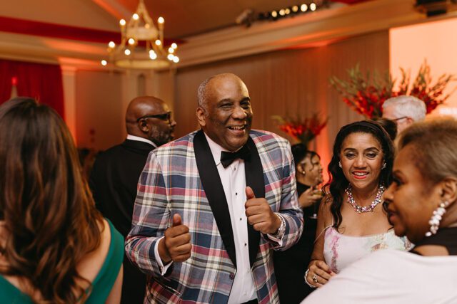 Man in plaid jacket and bow tie smiles, giving thumbs up.