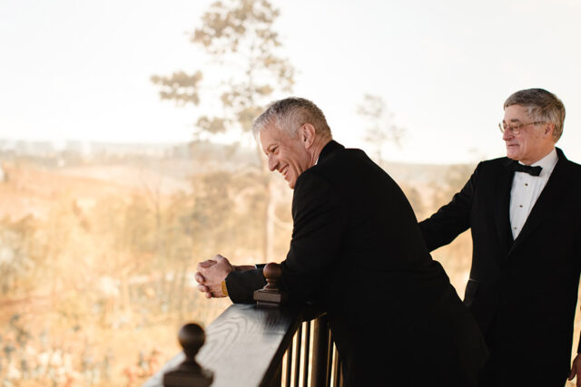 Two men in tuxedos stand on a balcony, one laughing and leaning on the railing.
