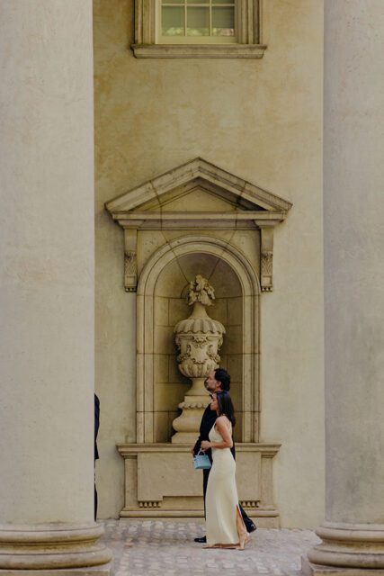 Couple in formal wear walking past a stone urn niche and columns