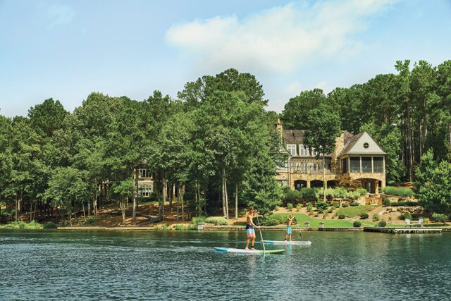 Two men paddleboarding on a lake in front of a large stone house surrounded by trees.