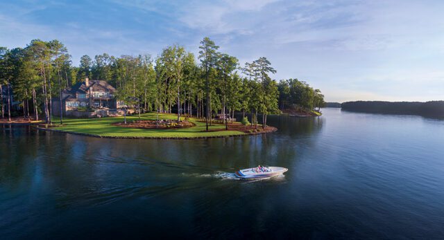 Luxury lake house with boat on water, surrounded by trees and manicured lawn.