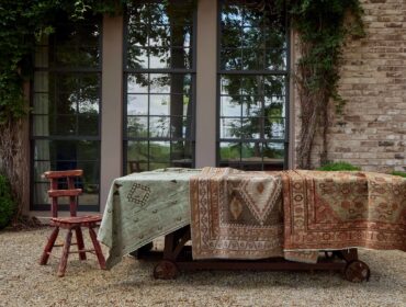 Brick building exterior with large windows, ivy, and display of antique rugs on a cart.