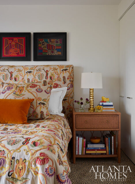 Bedroom with colorful embroidered bedding, orange pillow, and wicker nightstand with books and lamp.