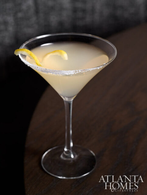 Cocktail in a martini glass with a sugar rim and lemon twist, on a dark wood surface.