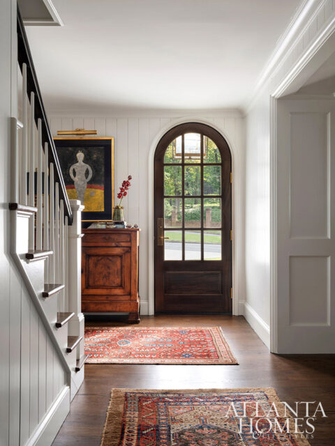 Entryway with arched wooden door, staircase, antique cabinet, and oriental rugs.