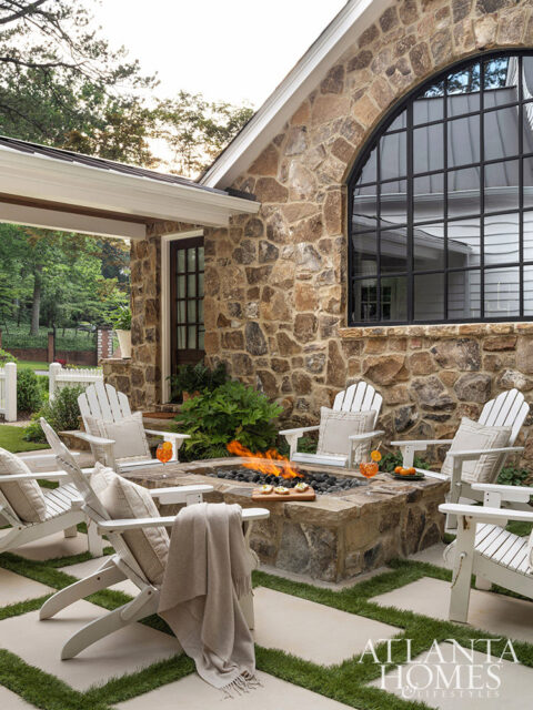 Stone house exterior with arched window, fire pit, and white Adirondack chairs.