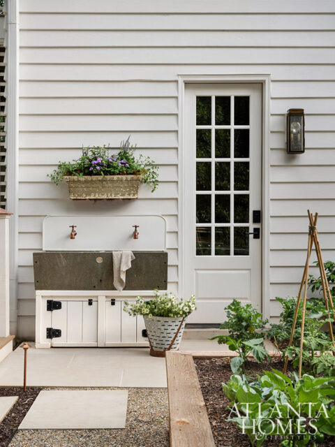 White paneled exterior with a glass-paned door, outdoor sink, and garden beds.