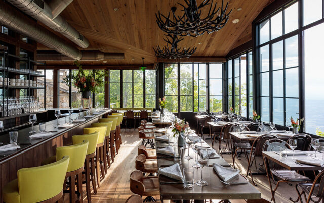 Restaurant dining room with bar, tables set for dinner, and large windows overlooking a view.