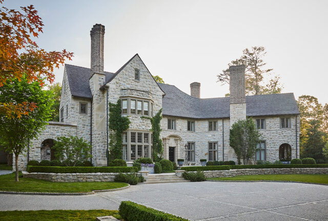 Stone mansion with multiple chimneys, arched entryway, and manicured hedges.