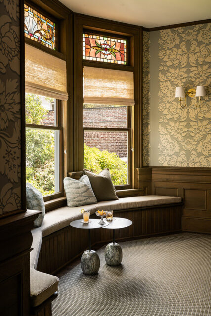 Window seat with stained glass, patterned wallpaper, and two small tables with drinks.