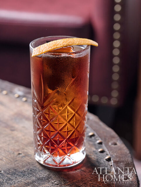 Tall glass of amber cocktail with ice and orange peel garnish on a wooden surface.