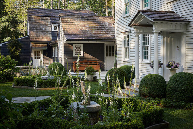 Shingled house with white columns and porch, surrounded by manicured gardens and foxglove flowers.