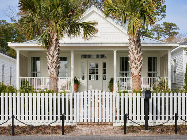 White house with a porch, palm trees, and a white picket fence.