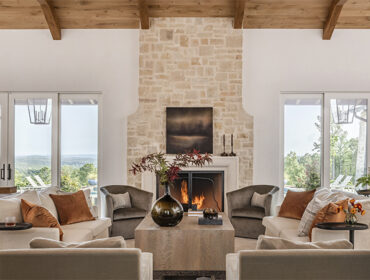 Living room with stone fireplace, wood beams, and neutral sofas with orange accents.