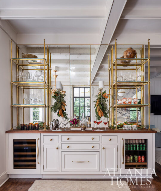 White bar with gold shelving, mirrored backsplash, and wine fridge stocked with drinks.