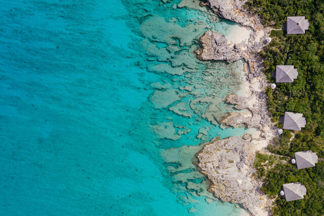 Aerial view of turquoise water, rocky coastline, and several grey-roofed pavilions nestled in lush