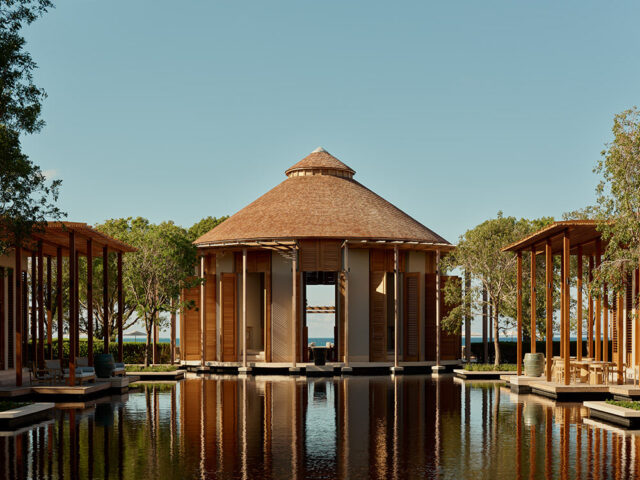 Resort arrival pavilion with wooden shutters and a thatched roof, reflected in a water feature.