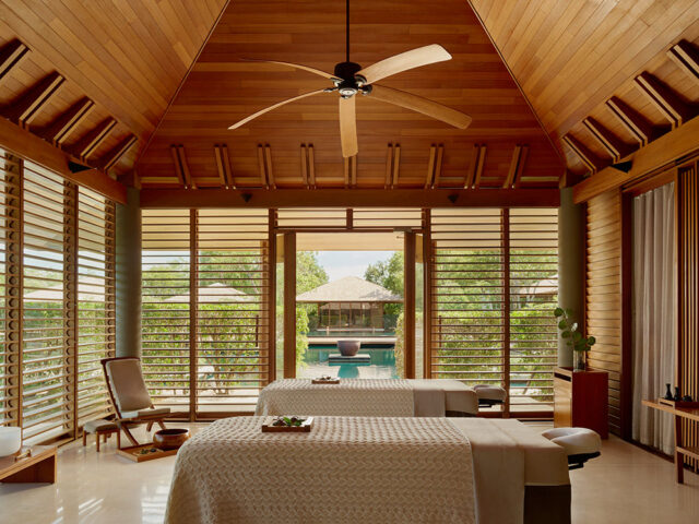 Spa treatment room with two massage tables, wood-paneled ceiling, and slatted windows overlooking a