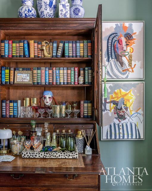 Wooden secretary desk with open hutch filled with books and barware. Two abstract portraits hang on