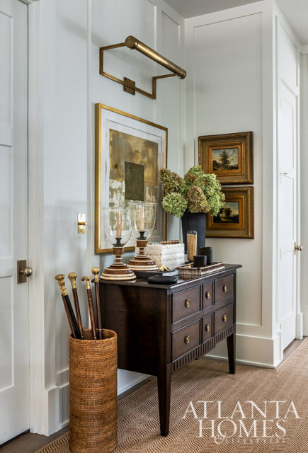 Hallway with dark wood chest, brass wall sconce, abstract art, and walking sticks in a basket.