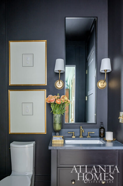 Dark powder room with gold-framed art, brass fixtures, and a vase of flowers.