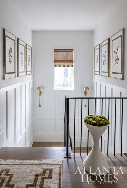 Hallway with white paneled walls, botanical prints, brass sconces, and a modern planter with moss.