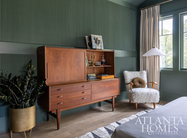 Green paneled walls with a mid-century modern teak secretary desk and a fluffy white armchair.