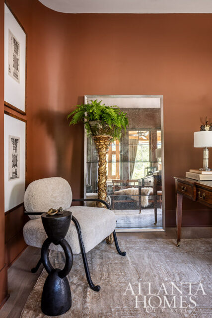 Living room with rust-colored walls, a textured cream armchair, ornate gold pedestal with fern, and